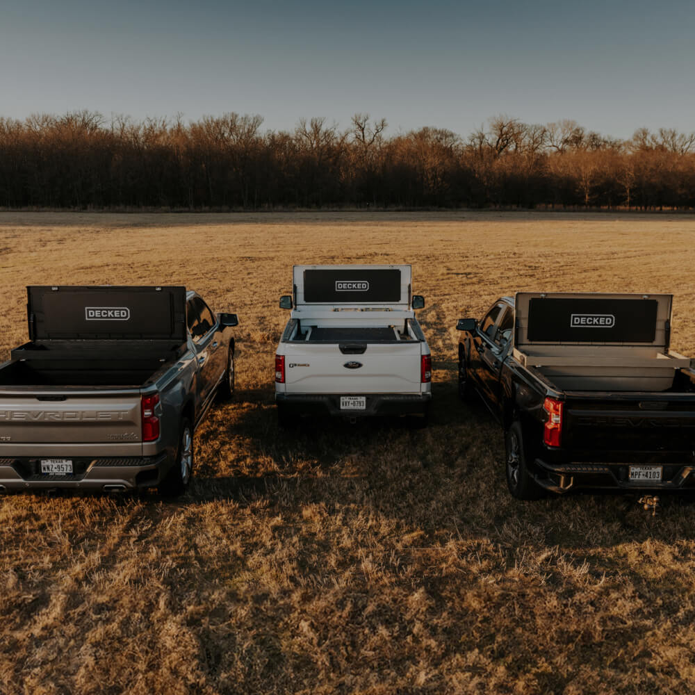 Three open DECKED Tool Boxes in the back of three truck beds in a grass field. 