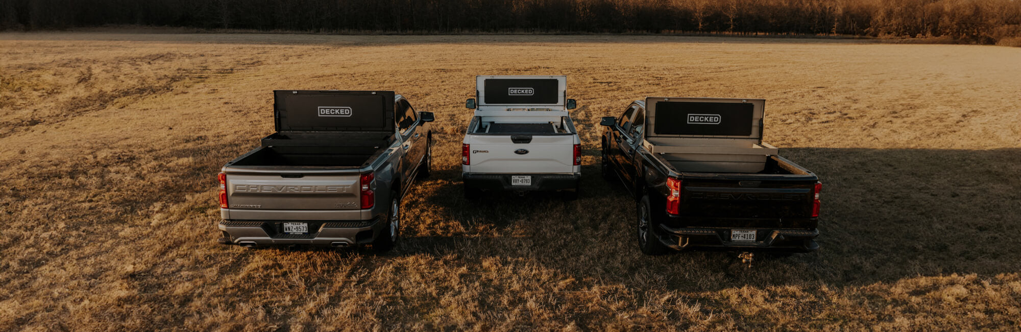 Three open DECKED Tool Boxes in the back of three truck beds in a grass field. 