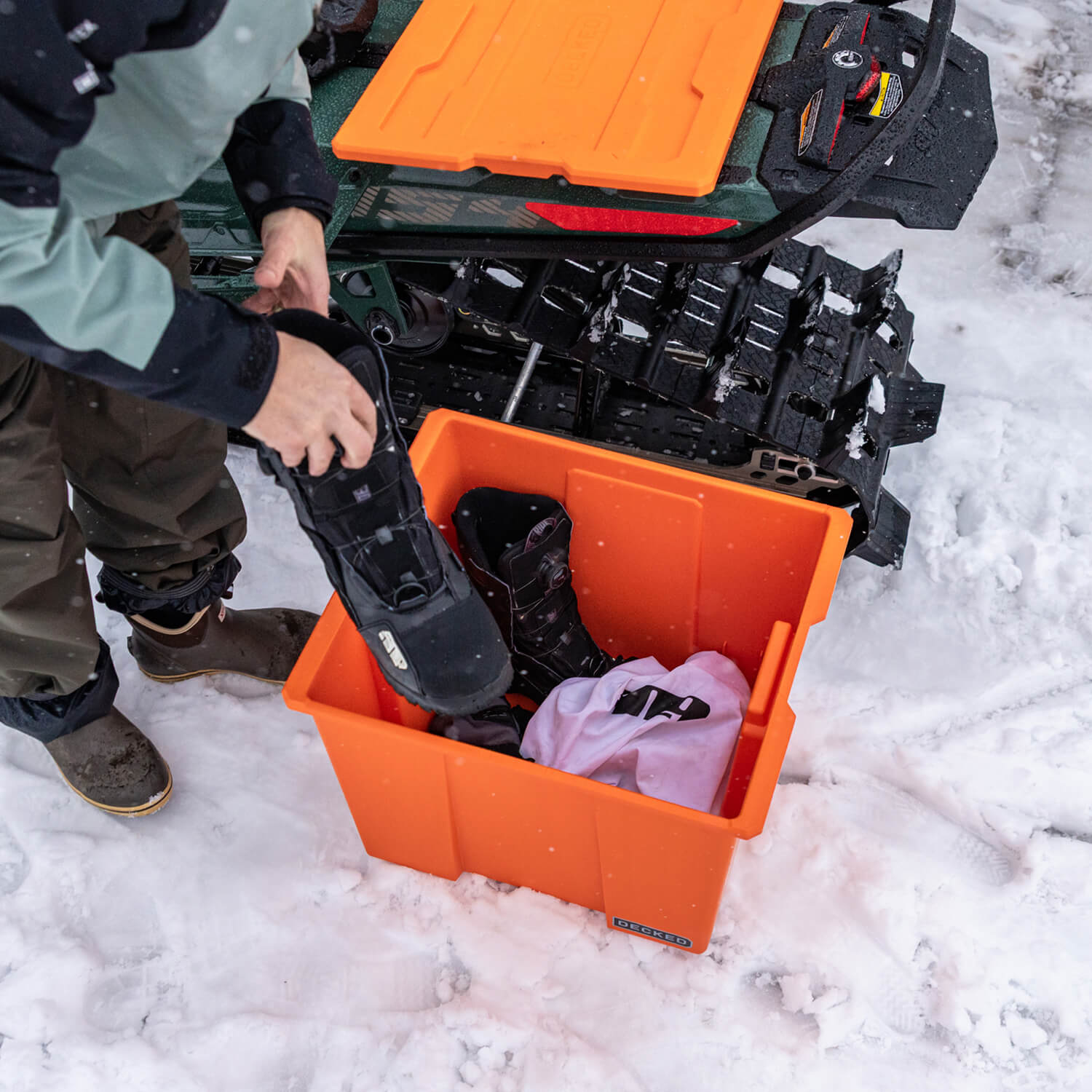 A blaze orange Payloader 64 sitting in the snow with snowmobiling boots and other gear inside.
