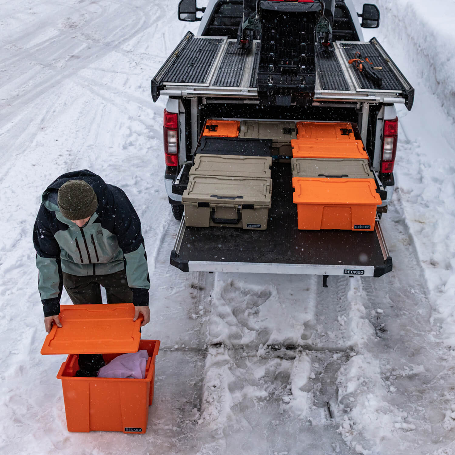 A person putting a lid on his blaze orange Payloader 64 bin next to his truck that has a CargoGlide, several protective cases and weatherproof bins, and a snowmobile loaded on the sled deck of the truck.