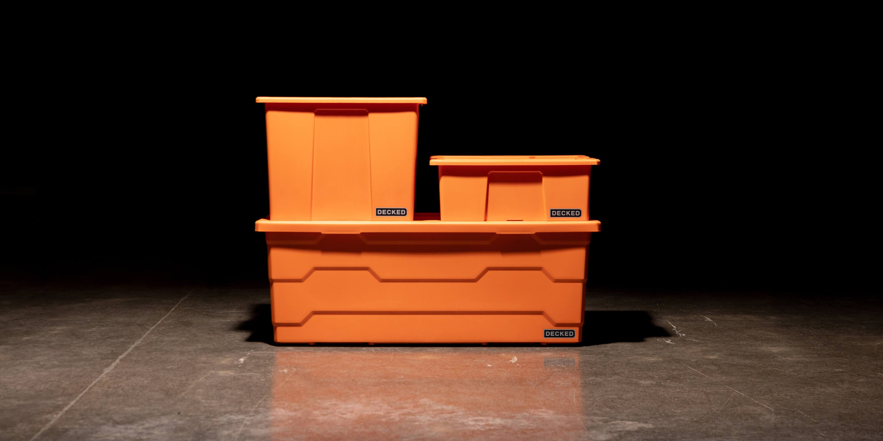 Three blaze orange Payloader Bin sizes stacked on top of each other in a garage with a dark, shadowy background.