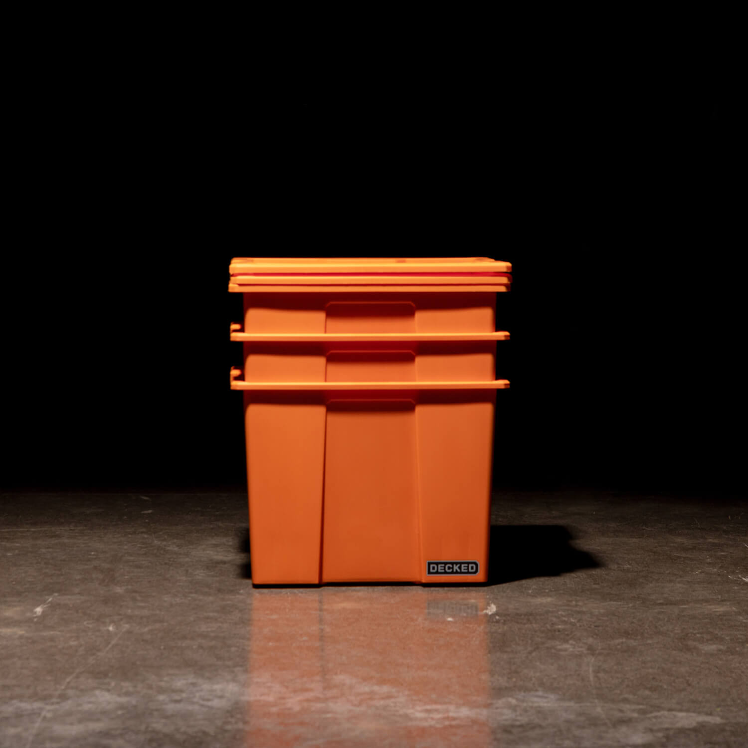 Three blaze orange Payloader 64 Bins nested together with the lids sitting on top in a garage with a dark, shadowy background.