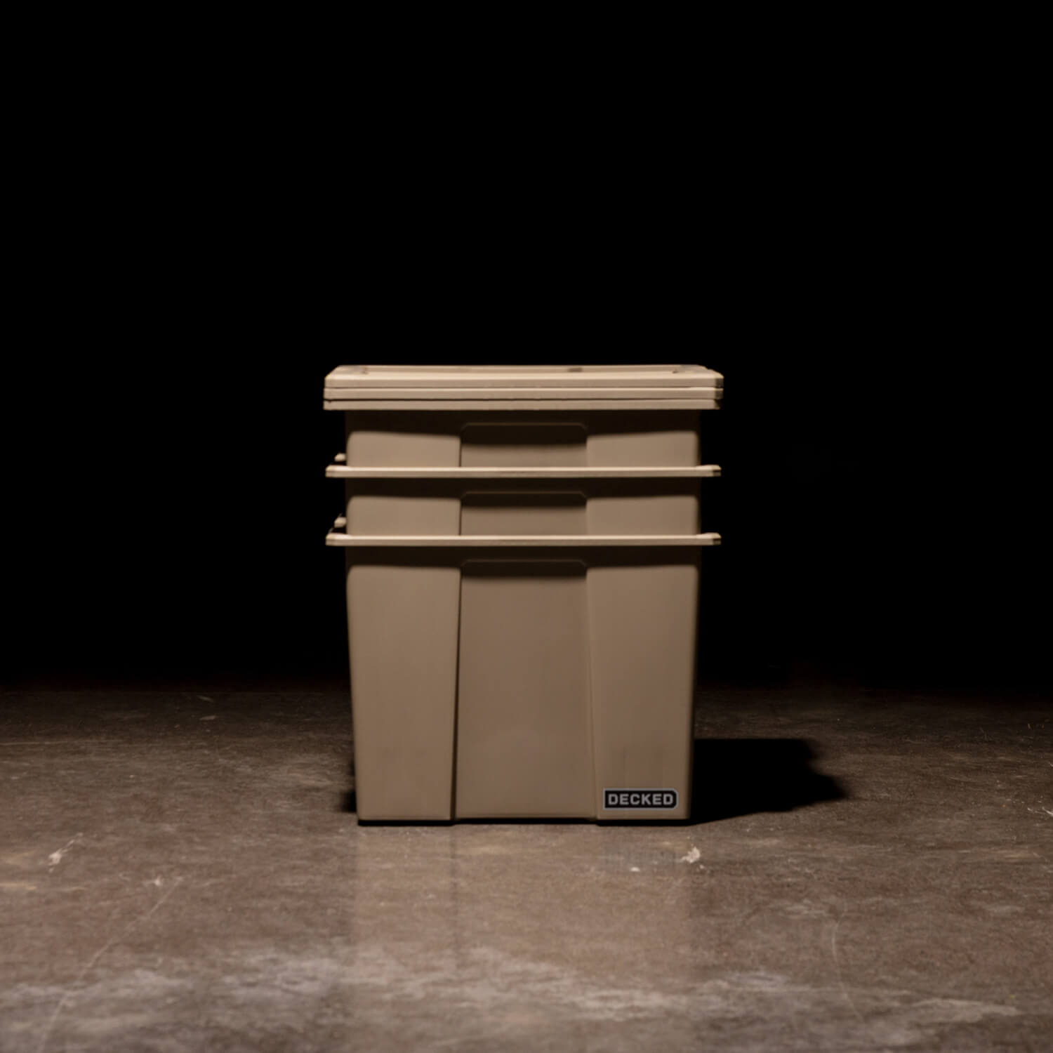 Three desert tan Payloader 64 Bins nested together with the lids sitting on top in a garage with a dark, shadowy background.