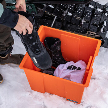 Person placing a snow boot into an orange Payloader 64 storage bin filled with items on a snowy ground.
