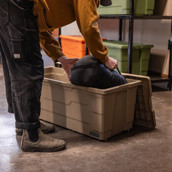 Person using a large Desert Tan Payloader 133 storage container with a lid, surrounded by shelves with green and orange boxes.