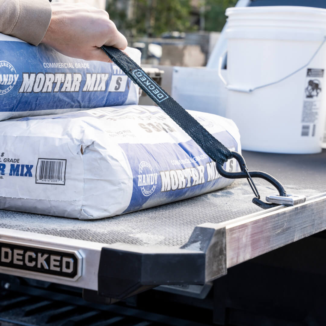 A DECKED cam strap attached to the D-rings for CargoGlide accessory holding down 80lb bags of mortar mix on top of a CargoGlide.