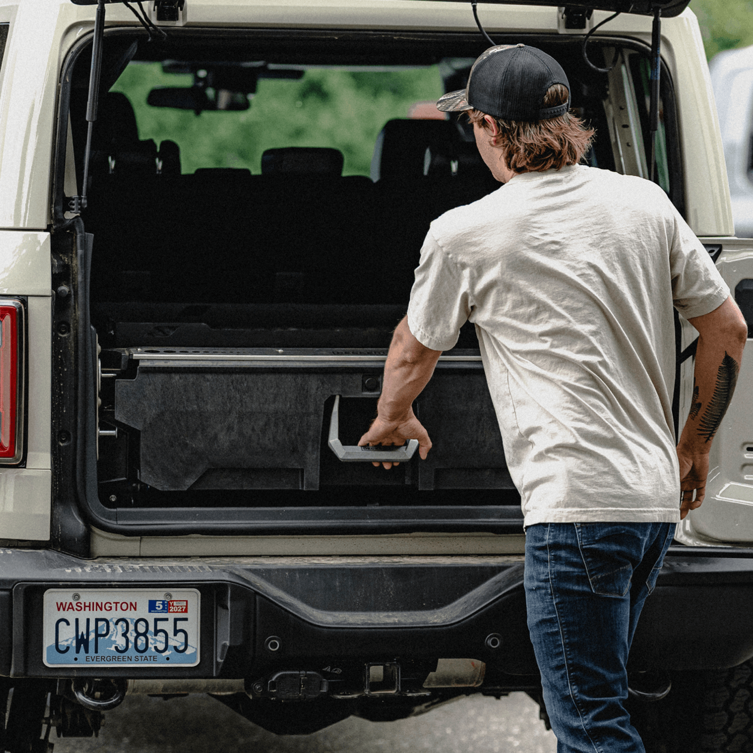 A man in a beige t-shirt and jeans stands at the open trunk of a white vehicle, pulling the drawer of a DECKED Drawer System open.