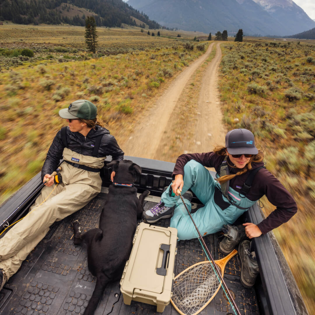 Two women sitting in the back of a truck with fly fishing rods and a sixer 16 between them