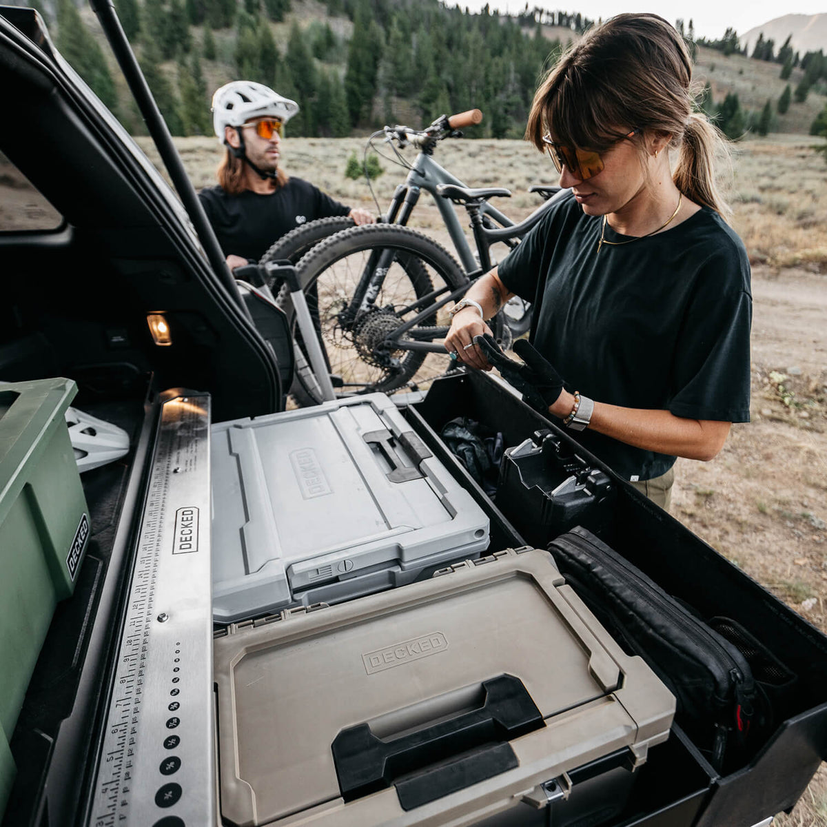 A woman and a man load gear into the back of their SUV with bicycles beside them, outdoors in a mountain setting. The foreground shows rugged hard cases and storage bins in the truck bed, including visible DECKED branding.