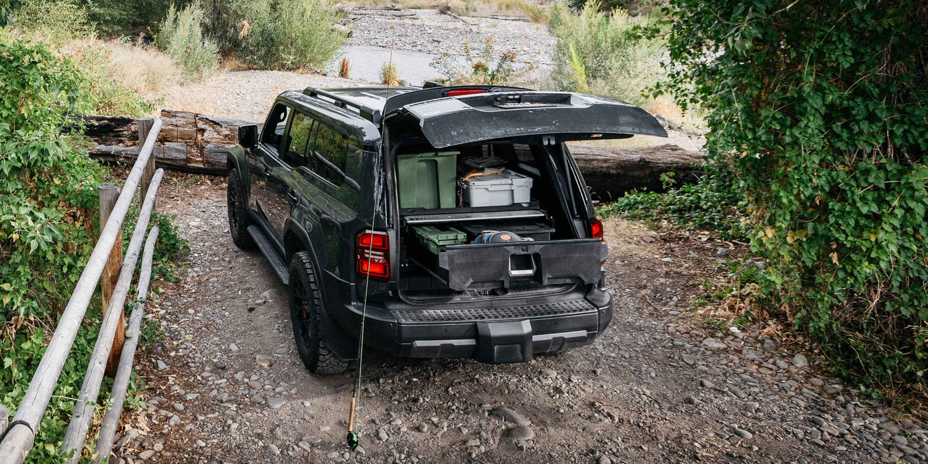 A Toyota Land Cruiser with the tailgate open near a river access. Inside the vehicle is a DECKED Drawer System and several rugged bins and fishing equipment and leaning against the car is a fly fishing rod.