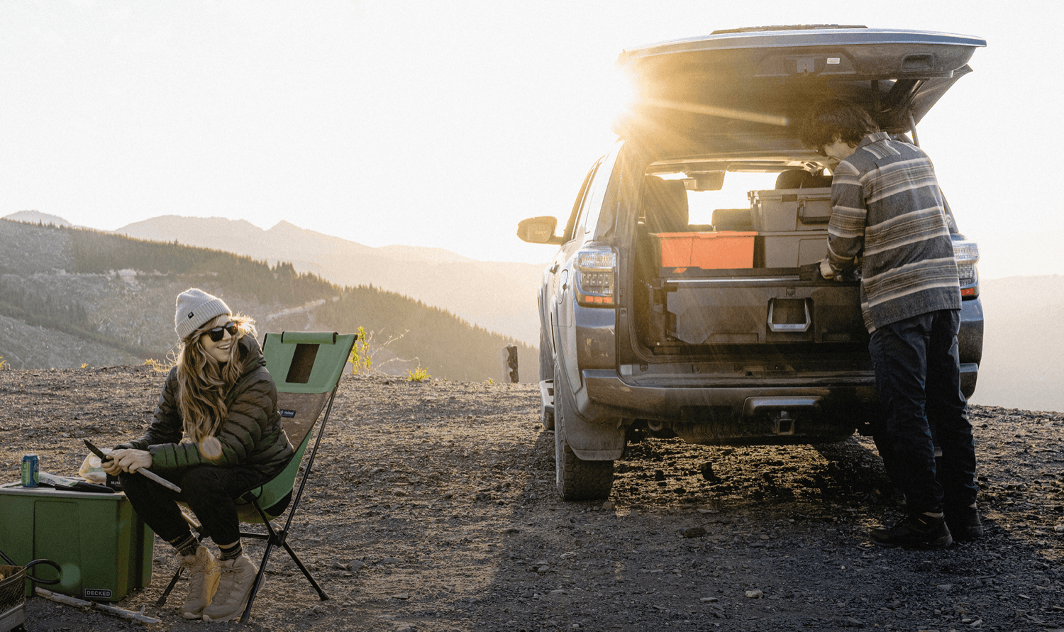 A lifestyle outdoor scene at sunset showing a woman sitting in a folding chair on the left and a man loading gear into his DECKED drawer system installed in the back of his Toyota 4Runner. 