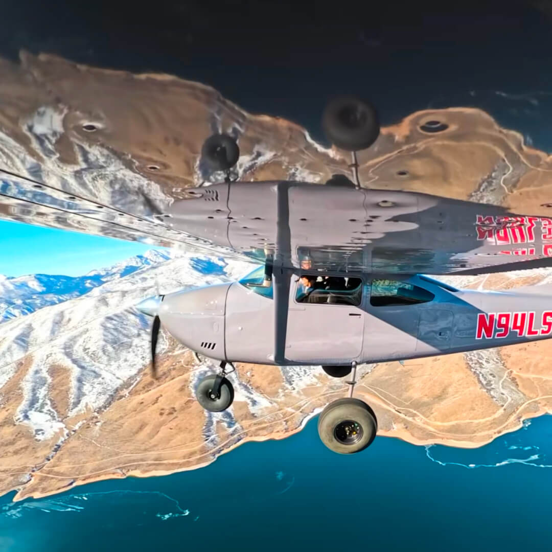 A shot of Chris flying a small plane, the mountains and water below are reflecting off of the underside of the wing.