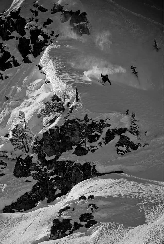 Snowboarder, Curtis Ciszek, turning on a snowy cliff with trees and mountains in the background