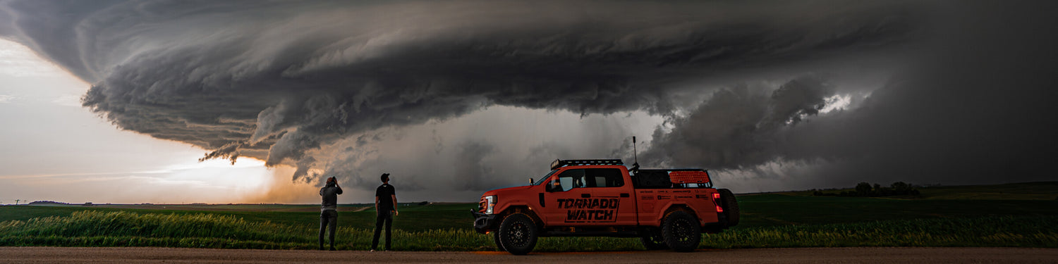 Ricky Forbes and his orange storm chasing rig, the Tornado Watch, on the side of a dirt road with what looks like a tornado building in the distance.