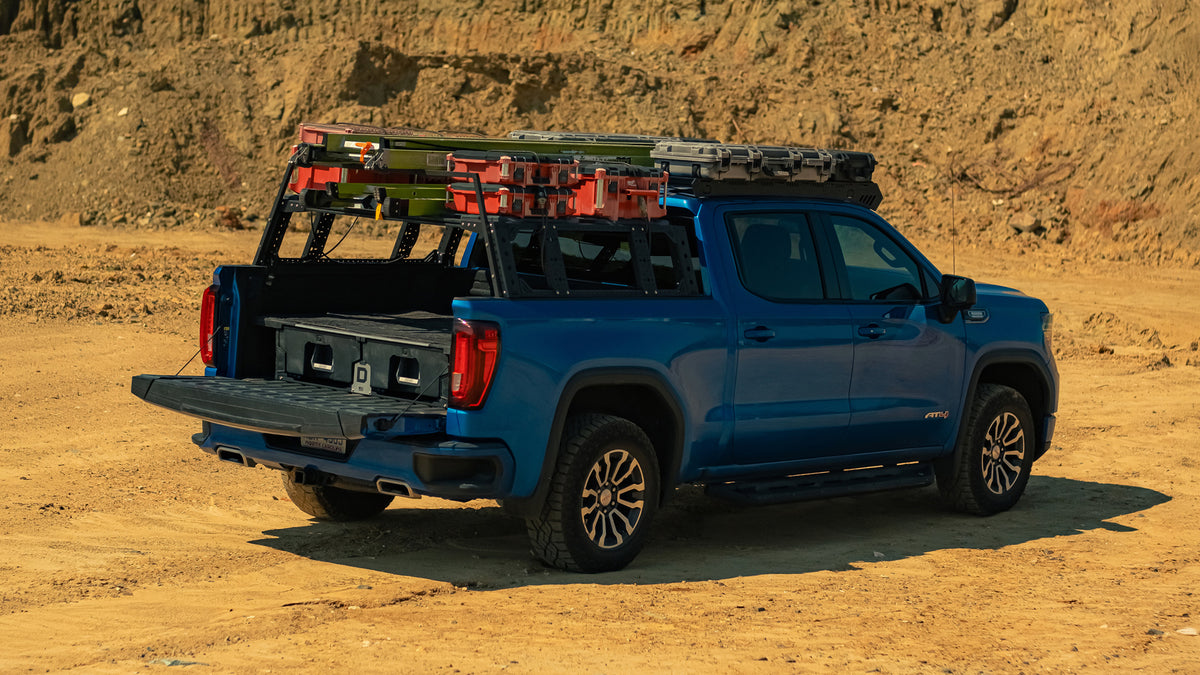 Thor Rippey’s blue pickup truck parked on a dirt lot with its tailgate down, showing a DECKED drawer system and ladders secured to the rack above the bed.