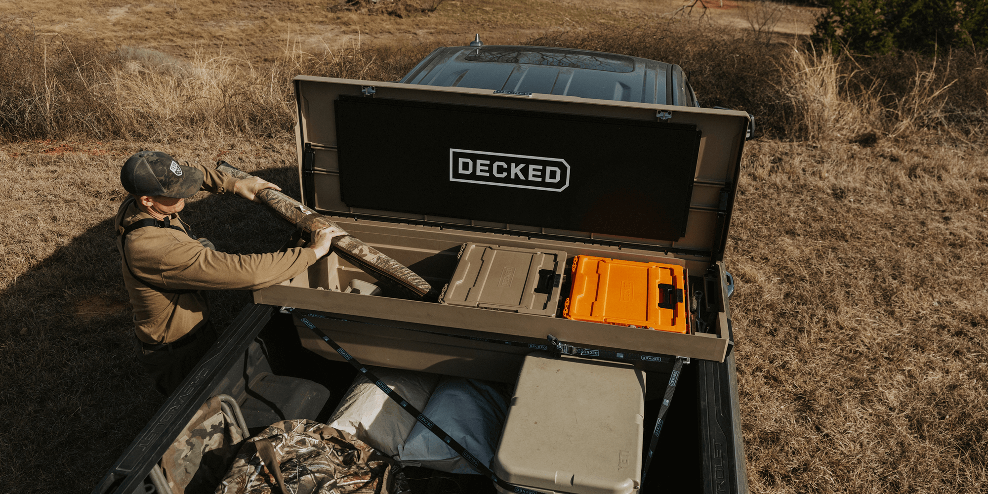 Person loading a shotgun case into their 'DECKED' Tool Box storage system in an outdoor setting.