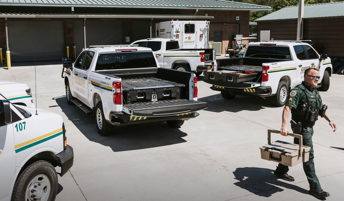 A sheriff walking with a Halfrack D-co case toolbox near a line of law enforcement trucks in a parking lot.