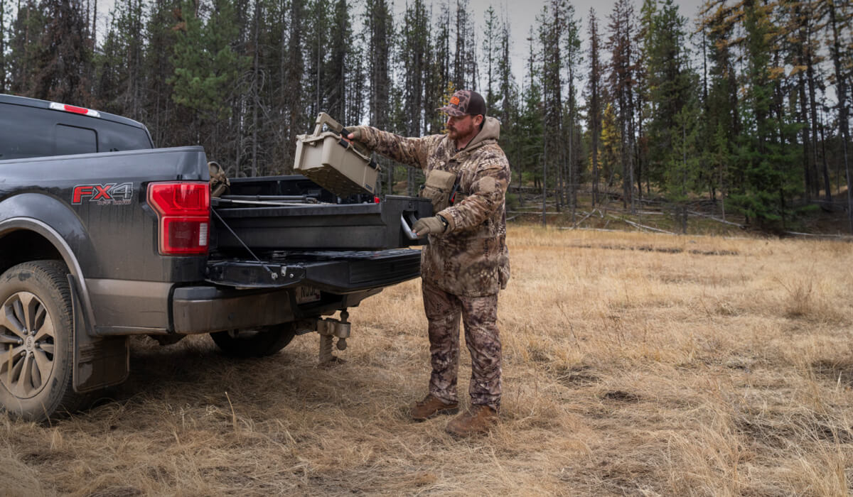 Man in camouflage gear loading a Sixer D-co case into the back of his pickup truck's Drawer System in a forested area.