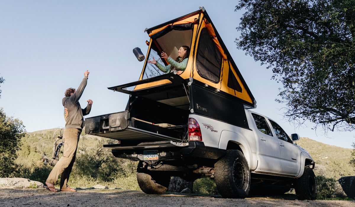 Two people interacting with a truck bed camper in a natural setting.
