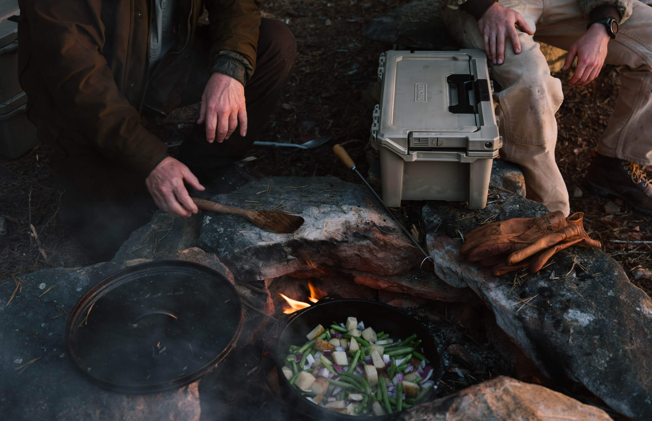 Person cooking outdoors over a fire pit with a Sixer D-co case holding their supplies nearby