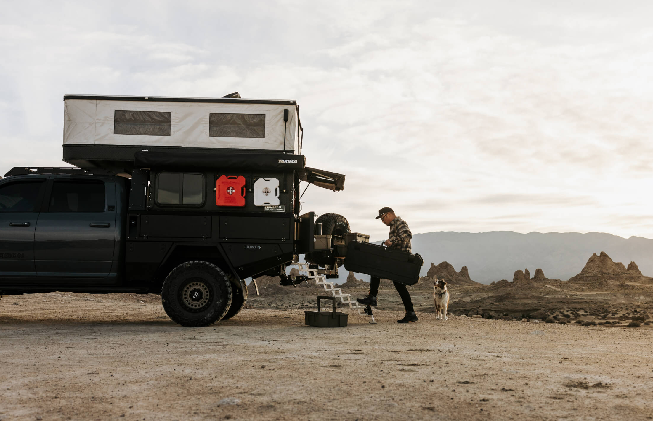 Person with a dog loading D-co cases into a black truck with a rooftop camper in a desert landscape.