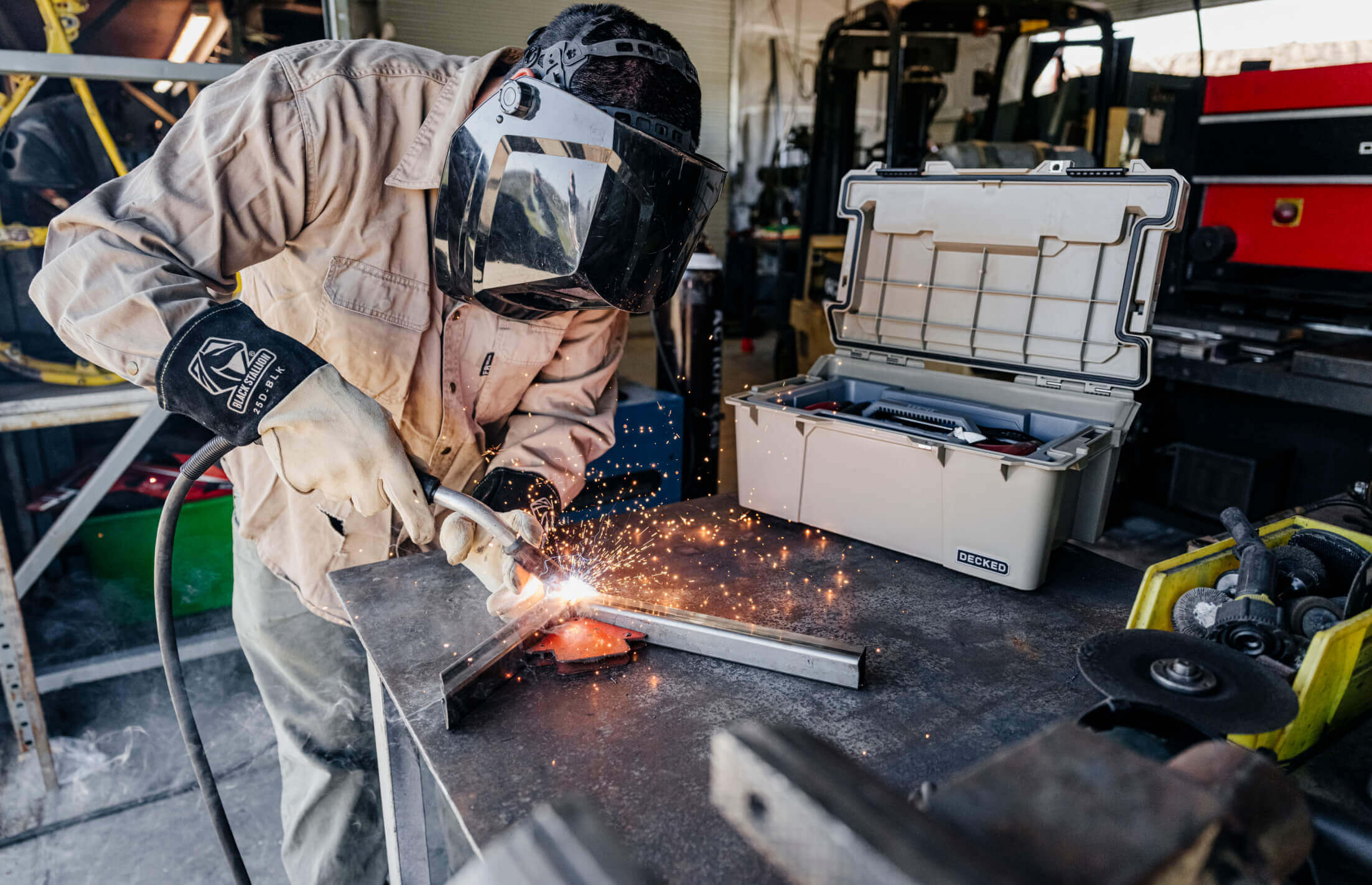 Person welding metal in a workshop with sparks flying and a Sixer D-co case toolbox sitting nearby.
