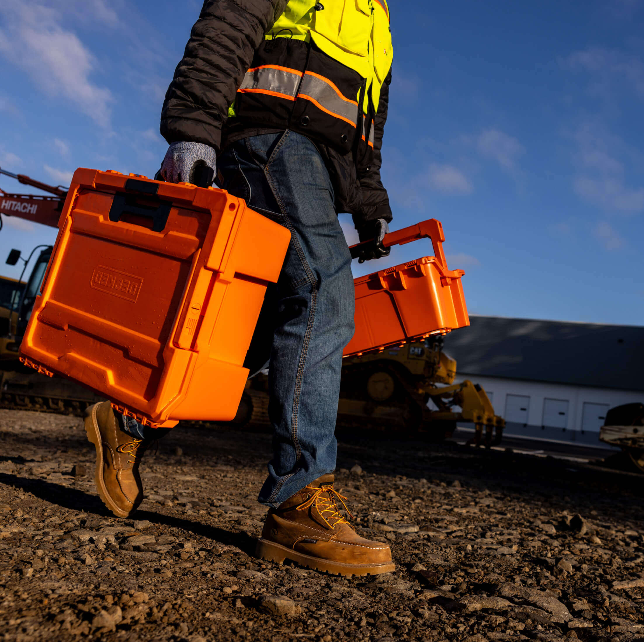 Person in high-visibility jacket carrying two orange D-co case toolboxes on a construction site