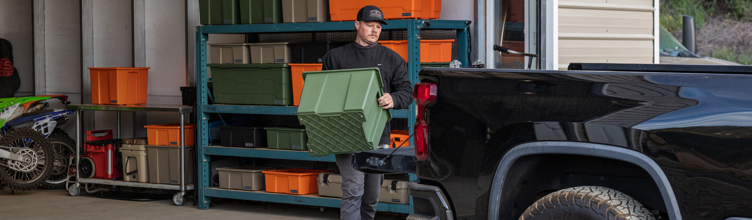 Person loading a green Payloader 64 storage bin onto a truck next to a storage rack with various bins and cases.