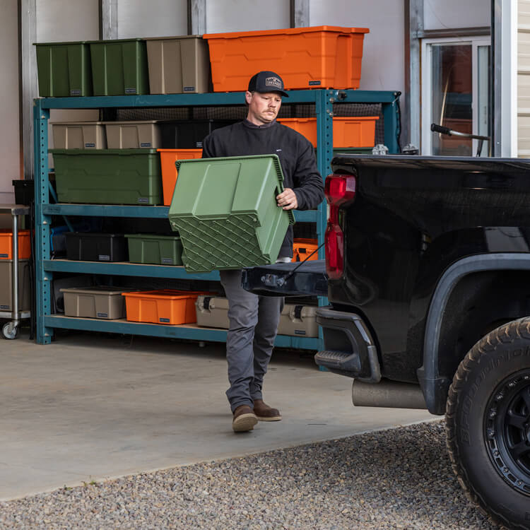 Person loading a green Payloader 64 storage bin onto a truck next to a storage rack with various bins and cases.