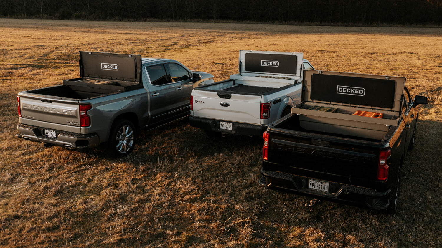 Three open tool boxes in the back of three truck beds in a grass field. 